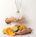 Eleganter Angel Wings Raumduft-Diffuser in cleanem Weiß mit silbernen Stäbchen im Torn-Paper-Look. Hände präsentieren den Flakon zusammen mit frischen Orangenscheiben, Bergamotte, Mango und Passionsfrucht durch weißes, gerissenes Papier – Best Bougie sanfter Premium Wohnduft.