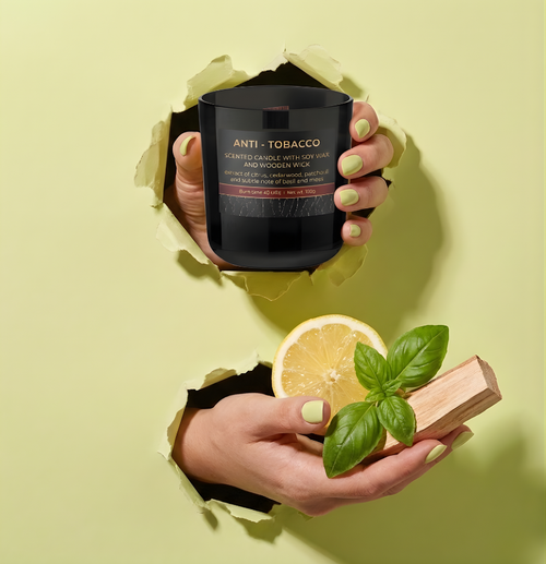 Hochwertige Anti-Tobacco Wood Duftkerze mit Sojawachs und Holzdocht im Torn-Paper-Look. Hände präsentieren die schwarze Kerze zusammen mit einer frischen Zitronenscheibe, Basilikumblättern und Zedernholz durch hellgrünes, gerissenes Papier – Best Bougie effektive Geruchsneutralisierung.