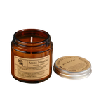 Hochwertige Aroma Sensation Duftkerze im Braunglas mit silbernem Deckel. Hände präsentieren die Kerze zusammen mit einer prachtvollen roten Rose und frischen Rosenknospen im Torn-Paper-Look durch ockerfarbenes Papier – Best Bougie floraler Premium Wohnduft.