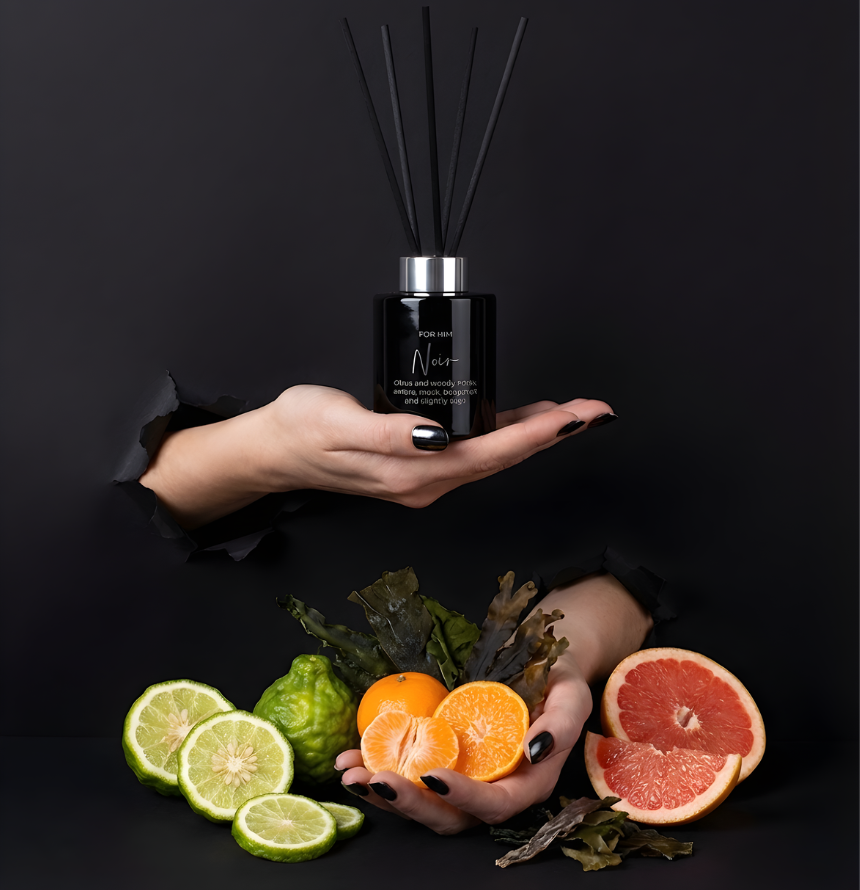 Eleganter Noir Raumduft-Diffuser „For Him“ in edlem Schwarz mit schwarzen Stäbchen im Torn-Paper-Look. Hände präsentieren den Flakon zusammen mit frischen Bergamotten, Mandarinen, Grapefruit-Hälften und dunklen Algenblättern vor schwarzem Hintergrund – Best Bougie maskuliner Premium Wohnduft.