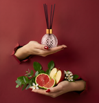 Langanhaltender Raumduft-Diffuser in einem rosa Kristall-Glasflakon mit schwarzen Stäbchen. Hände präsentieren den Diffuser zusammen mit Apfel, Grapefruit und Eukalyptus im Torn-Paper-Look durch dunkelrotes Papier – Best Bougie Premium Wohnduft.