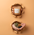 Hochwertige Duftkerze God’s SPA Harmony im Torn-Paper-Look. Hände präsentieren die Kerze zusammen mit getrockneten Patchouli-Blättern, Ambra-Harz und Tonkabohnen durch pfirsichfarbenes, gerissenes Papier – Best Bougie harmonischer Raumduft.