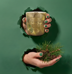 Hochwertige Duftkerze Golden Ginkgo mit Sojawachs und Holzdocht im Torn-Paper-Look. Hände präsentieren die Kerze mit goldenem Ginkgo-Dekor zusammen mit frischen Kiefernzweigen und Zapfen durch grünes, gerissenes Papier – Best Bougie naturnaher Raumduft.