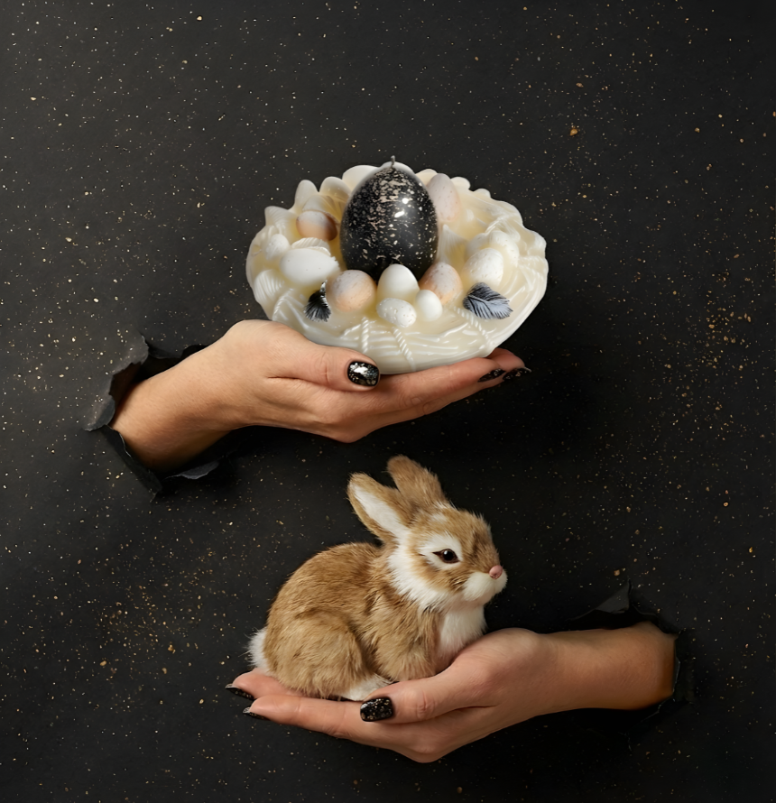 Kreative Oster-Kerze in Nestform mit schwarz-golden gesprenkeltem Ei im Torn-Paper-Look. Hände präsentieren das Kerzen-Nest zusammen mit einem kleinen, flauschigen Osterhasen durch schwarzes, mit Goldstaub besprenkeltes Papier – Best Bougie exklusive Deko-Duftkerze für Ostern.