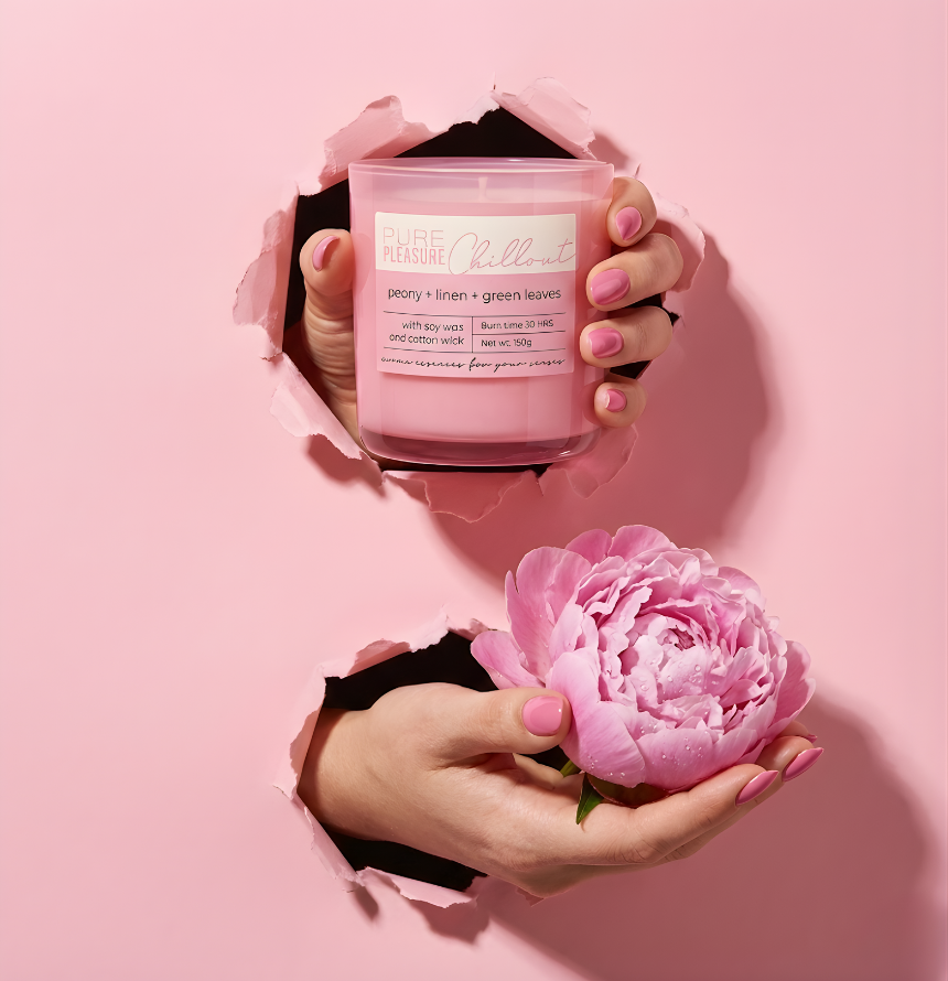 Stilvolle Präsentation der floralen Duftkerze Pure Pleasure Chillout. Hände ragen durch pinkes, zerrissenes Papier und halten die Kerze neben einer großen, frischen Pfingstrose – Best Bougie Premium Raumduft für Frauen