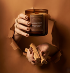 Stilvolle Präsentation der hochwertigen Duftkerze Sandalwood mit Holzdocht. Hände reichen durch einen warmen, erdfarbenen Hintergrund und halten die Kerze sowie rohe Sandelholz-Stücke – Best Bougie natürliche Dufterlebnisse